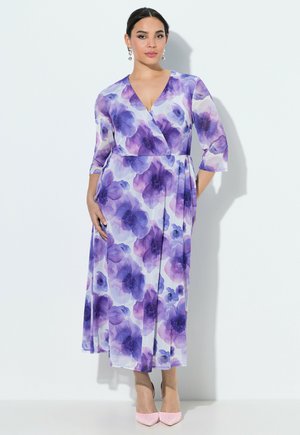 Mujer de pie vistiendo un vestido largo cruzado con estampado floral en púrpura y blanco, con mangas de tres cuartos y tacones puntiagudos rosas.