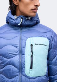 Homme portant une veste matelassée bleue avec une capuche et une poche poitrine zippée bleu clair étiquetée « PeakPerformance ».