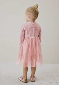 Vaaleanpunainen mekko, jossa on pitsineulottu yläosa pitkähihaisilla hihoilla, röyhelöinen tüllihame ja koristeelliset napit takana; yhdistettyinä vaaleanpunaisiin kenkiin.