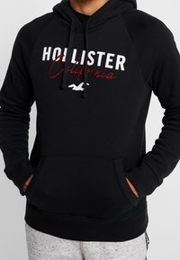 Svart huvtröja i mjukt tyg, med känguruficka, snodd i huvan och broderad "Hollister California"-logotyp i vitt och vinrött.
