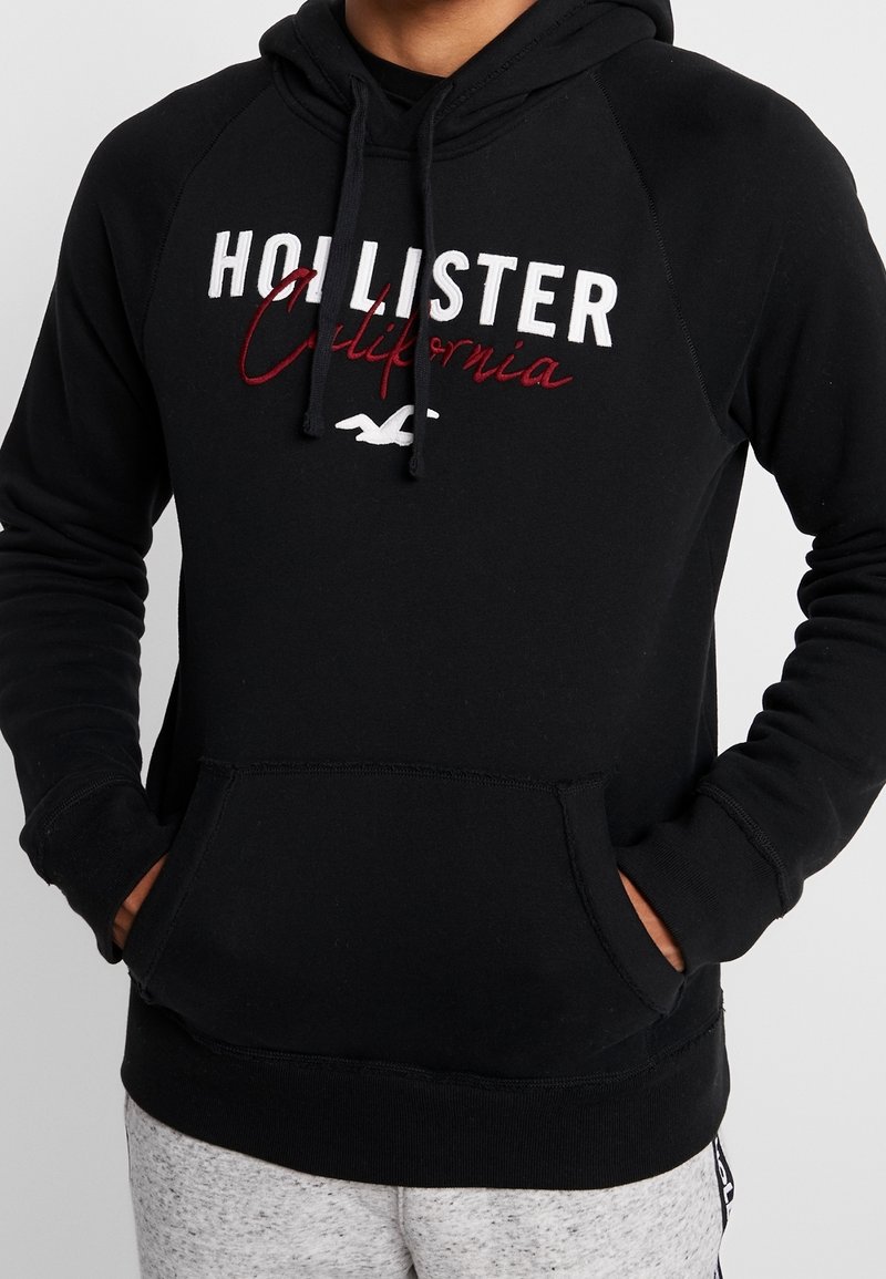 Svart huvtröja i mjukt tyg, med känguruficka, snodd i huvan och broderad "Hollister California"-logotyp i vitt och vinrött.