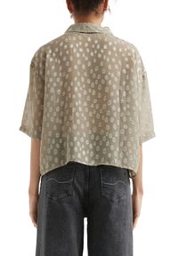 Blusa leggera e trasparente di colore verde oliva con una vestibilità morbida, caratterizzata da un motivo a pois in tonalità più chiare. Maniche corte e colletto.