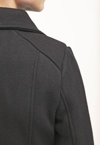 Henkilö, joka on pukeutunut mustaan villatakkiin, kuvattu yläselästä ja olkapäästä vaaleataustaista taustaa vasten.