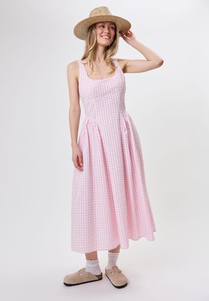 Kobieta ubrana w różowo-białą sukienkę w kratkę bez rękawów, beżowe chodaki z skarpetkami oraz słomkowy kapelusz z szerokim rondem, uśmiechająca się i dotykająca kapelusza.