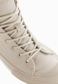 Zapatilla alta beige con parte superior de cuero suave, cordones planos y suela de goma con puntera texturizada y ojales de metal.