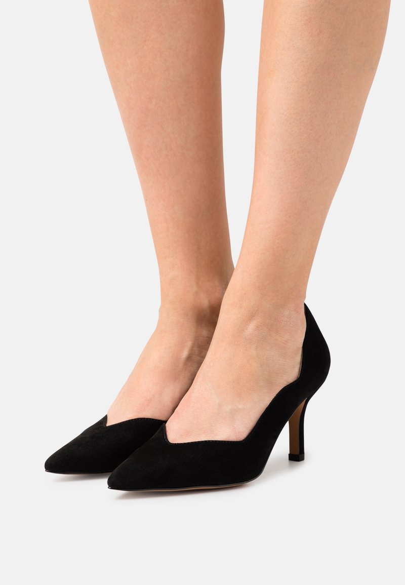 Jambes féminines portant des escarpins en daim noir à bout pointu et talon moyen sur un fond clair uni.
