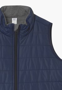 Granatowa pikowana kamizelka puchowa z wysokim kołnierzem, zamkiem błyskawicznym z przodu i szarym podszyciem. Bezrękawnik o miękkiej fakturze.