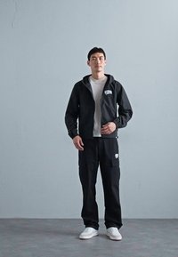 Giacca leggera nera con zip e cappuccio, abbinata a pantaloni cargo neri con tasca a toppa, e sneakers bianche. T-shirt bianca sotto.