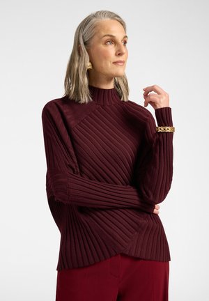 Frau mit grauen Haaren trägt einen gerippten burgunderfarbenen Pullover und passende Hose, accessoriert mit goldenem Armband und Ohrringen, schaut zur Seite.