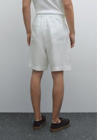 Shorts en lin blancs avec une coupe décontractée, taille élastique et deux poches arrière appliquées. Portés avec des chaussures foncées sur une surface gris texturé.