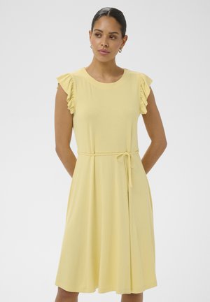 Femeie purtând o rochie galben pal până la genunchi, cu mâneci scurte cu volane și o curea legată în talie, stând pe un fundal simplu și deschis la culoare.