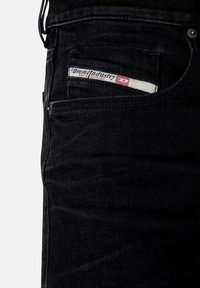 Jeans de mezclilla negra con una textura suave, que presentan una etiqueta de logo en el bolsillo frontal y sutiles detalles de costura. Diseño clásico de cinco bolsillos.