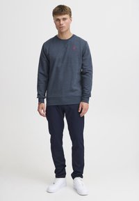 Sudadera azul marino con cuello redondo, mangas largas y un pequeño logo rojo en el pecho, combinada con pantalones azul marino y zapatillas blancas.