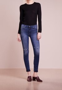 Vaqueros ajustados de color azul con detalles desgastados, combinados con una blusa negra de manga larga y unas mules de tacón morado oscuro, sobre un fondo neutro.