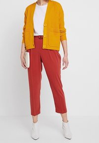 Žlutý žebrovaný kardigan s knoflíky vpředu a kapsami, bílé tričko, červené zúžené kalhoty s opaskem a bílé kotníkové boty.