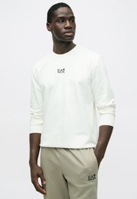 Cămașă off-white cu mâneci lungi, având un mic logo negru pe piept, asortată cu pantaloni jogger bej cu buzunare laterale și accente cu logo.