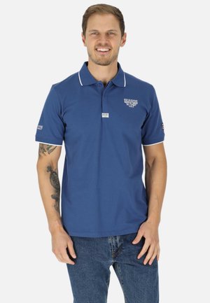 Uomo sorridente che indossa una polo blu con bordi bianchi e testo, abbinata a jeans blu, in piedi davanti a uno sfondo bianco semplice.