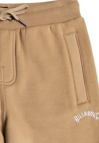 Tan-coloured sweatshortsit valmistettu pehmeästä kankaasta. Varustettu ribatusta vyötäröstä, jossa on kiristysnauha, sivutaskulla ja brodeeratulla "Billabong"-logolla.