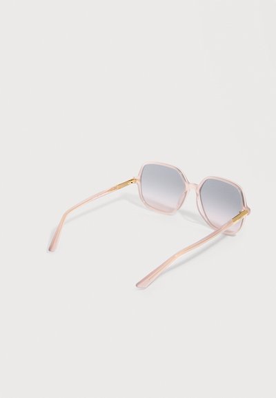 Lunettes de soleil à monture carrée avec branches translucides rose pâle et verres gris dégradés, posées ouvertes sur une surface blanche.