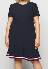 Persona con curvas vistiendo un vestido de manga corta, de color azul marino oscuro, con un dobladillo acampanado que presenta acentos a rayas rojas y blancas en la parte inferior.
