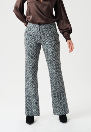 Woman wearing patterned wide-leg trousers and a brown long-sleeve blouse, standing against a plain light background.