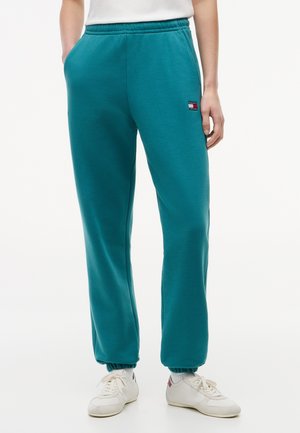 Άτομο που φοράει πετρόλ φόρμα με ελαστική μέση και ελαστικές μανσέτες, λευκά αθλητικά παπούτσια και λευκό τοπ, στέκεται με το ένα χέρι στην τσέπη.