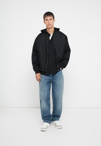 Chaqueta bomber negra con cierre frontal, llevada sobre una camisa negra de botones y jeans desgastados de un azul claro, complementada con zapatillas blancas.