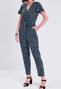 Combinaison verte et bleue à motifs avec un décolleté en V, des manches courtes, une taille cintrée et des jambes fuselées. Associée à des sandales à talons noires.