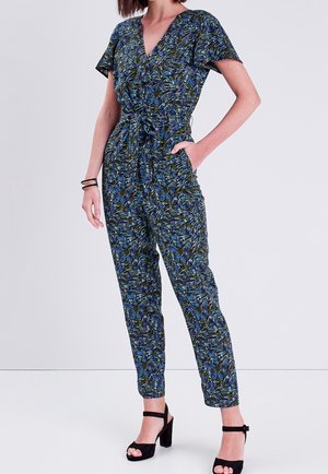 Combinaison verte et bleue à motifs avec un décolleté en V, des manches courtes, une taille cintrée et des jambes fuselées. Associée à des sandales à talons noires.