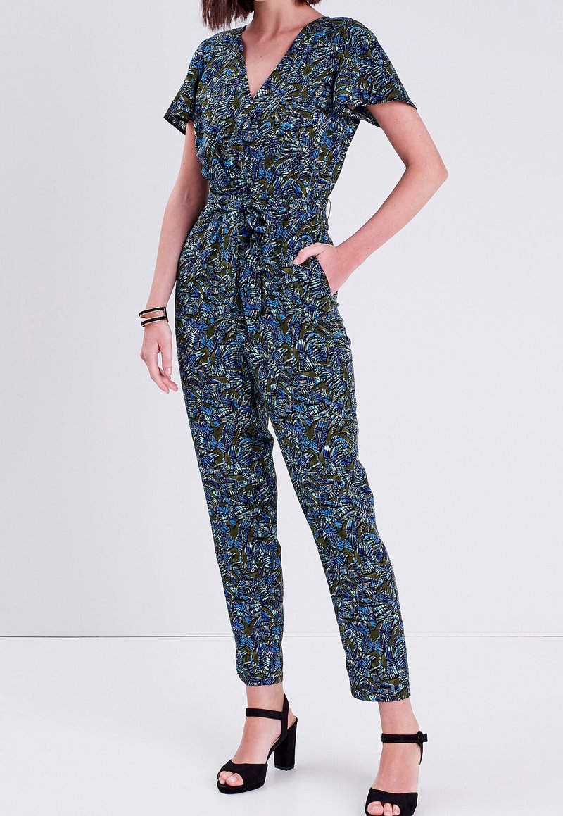 Combinaison verte et bleue à motifs avec un décolleté en V, des manches courtes, une taille cintrée et des jambes fuselées. Associée à des sandales à talons noires.