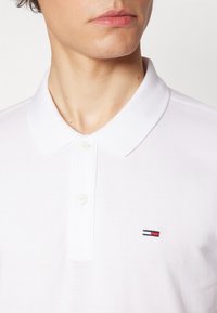 Camisa polo blanca hecha de tela texturizada, con un cuello clásico, tapeta de tres botones y un pequeño logo bordado en rojo, blanco y azul.