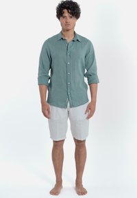 Lichtgroene linnen blouse met lange mouwen en knopen, gecombineerd met witte linnen shorts. Blootvoets in een stand tegen een witte achtergrond.