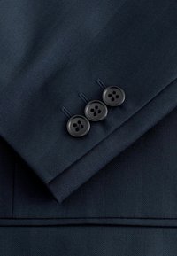 Manche de costume bleu marine foncé avec un motif chevron subtil et trois boutons noirs cousus en ligne près du poignet.