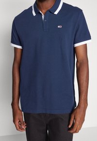 Polo bleu marine avec un col blanc, patte de boutonnage à deux boutons, et logo brodé sur la poitrine. Manches courtes avec un liseré blanc.
