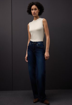 Hvid ærmeløs top med rund halsudskæring, kombineret med mørkeblå jeans med vide ben. Glat tekstur, diskrete syningsdetaljer og bæltestropper.