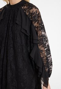 Schwarzes Spitzenkleid mit durchsichtigen, rüschierten Ärmeln, einem hohen Ausschnitt, strukturierten floralen Mustern und einer fließenden Silhouette.