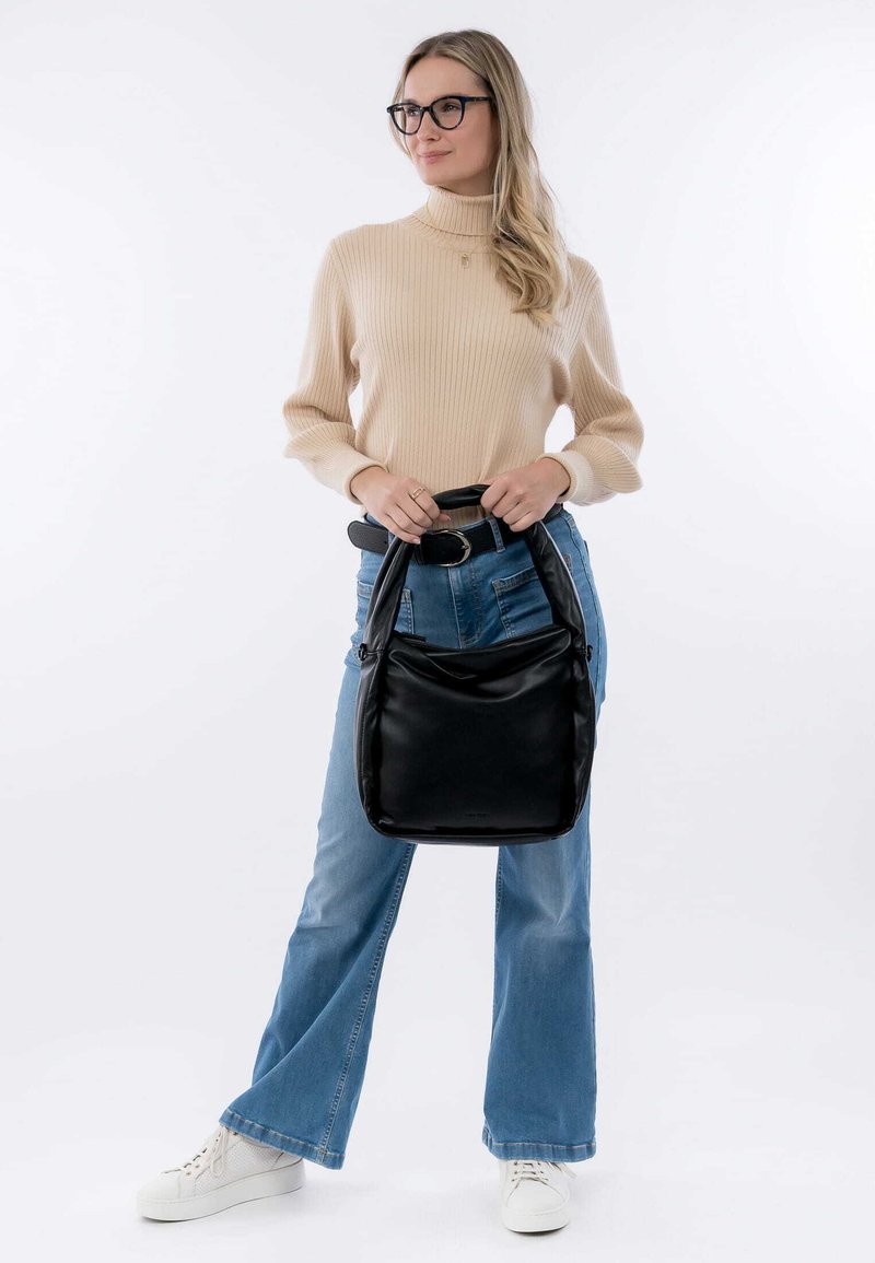 Borsa a tracolla in pelle nera con una forma arrotondata e una texture morbida, dotata di doppio manico. Modella che indossa un maglione beige a coste e jeans svasati blu.
