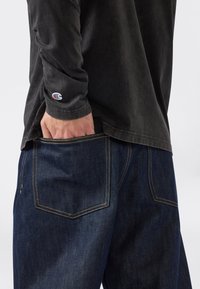 Person in dunklen Jeans mit der Hand in der Gesäßtasche und einem langärmeligen schwarzen Shirt mit einem Champion-Logo am Handgelenk.