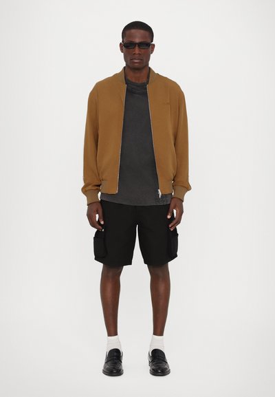 Veste bomber zippée marron sur une chemise grise, shorts cargo noirs et chaussures noires avec des chaussettes blanches, se tenant contre un fond blanc.