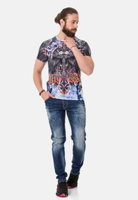 Schwarzes und blaues Grafik-T-Shirt mit kunstvollen Designs von Adler und Totenschädel; kombiniert mit zerrissenen blauen Jeans und schwarzen Sneakers mit roten Akzenten.