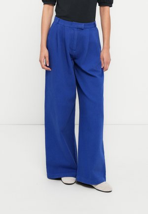 Weit geschnittene Hosen aus hellblauem Baumwollstoff. Verfügen über eine plissierte Vorderseite und eine gerade Passform. Kombiniert mit einem schwarzen Oberteil und grauen Schuhen.