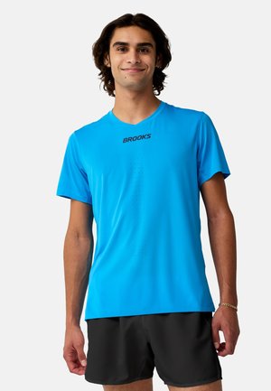 Jonge man met middellang donker haar, die een felblauw Brooks T-shirt en zwarte korte broek draagt, staand tegen een eenvoudige achtergrond.