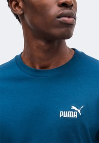 Άνδρας με μπλε T-shirt Puma με λευκό λογότυπο Puma στο στήθος, το κεφάλι γυρισμένο ελαφρώς δεξιά.