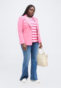 Γυναίκα με πλεξούδες, φορώντας ροζ σακάκι, ριγέ ροζ και λευκό πουκάμισο, μπλε καμπάνες τζιν, λευκά αθλητικά παπούτσια, κρατώντας μία μπεζ τσάντα.