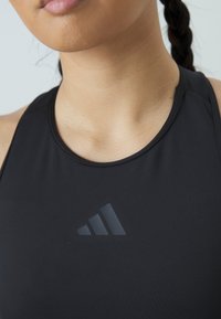 Crni sportski top bez rukava s okruglim izrezom, koji se ističe uzdignutim crnim logotipom na prsima i glatkom, rastezljivom tkaninom.