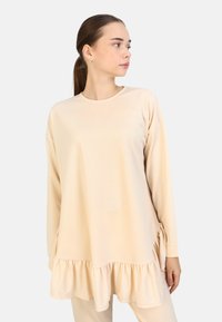 Lange mouwen, lichtbeige top met een ronde hals en een ruchesdetail aan de onderkant. Glad stof met een losse pasvorm.