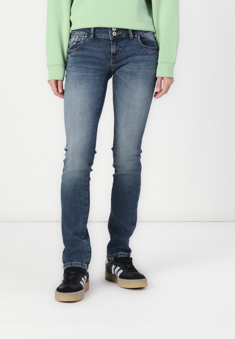 Jean bleu ajusté avec une texture délavée, présentant un design à cinq poches et une fermeture à deux boutons, assorti de baskets noires et blanches.
