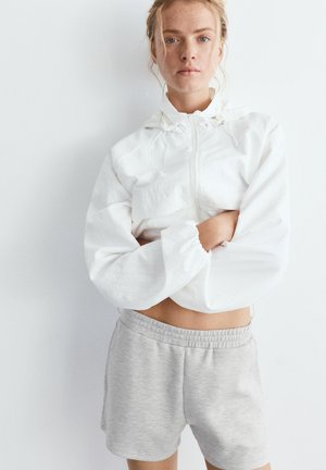 Fiatal nő szőke hajjal, fehér cipzáras rövid dzsekit és világosszürke rövidnadrágot visel, karját keresztbe téve áll, egyszerű háttér előtt.