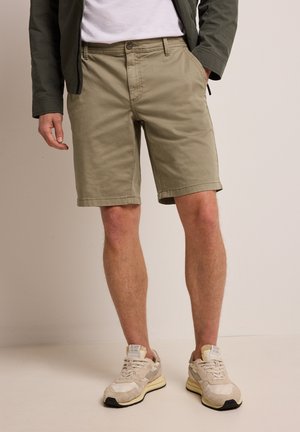 Homme portant un short beige mi-cuisses, des baskets blanches, une veste verte et un t-shirt blanc, debout avec une main dans la poche.
