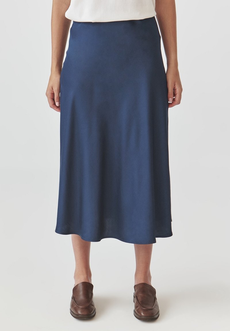 Femme portant une jupe bleu marine mi-genoux, un haut blanc et des chaussures marron sans lacets, debout devant un fond uni.